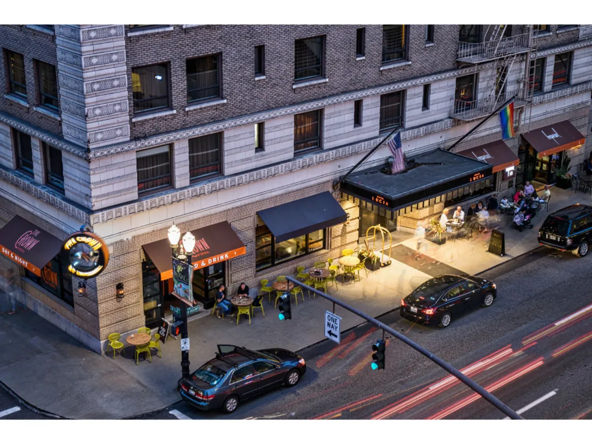 The photo captures a bustling outdoor restaurant scene with illuminated signage, people gathered at tables, and city lights reflecting the energy of the area. Moving cars and glowing traffic signals add to the dynamic ambiance of the city at night.