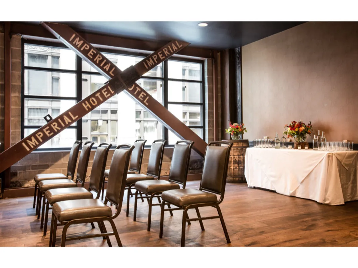 A spacious yet intimate room with rows of chairs, a decorative table adorned with flowers, expansive windows framing city views, and a historic 'Imperial Hotel' crossbeam sign overhead. The setting is elegant and versatile, hinting at the hotel’s storied past and its connection to Portland’s vibrant urban life.
