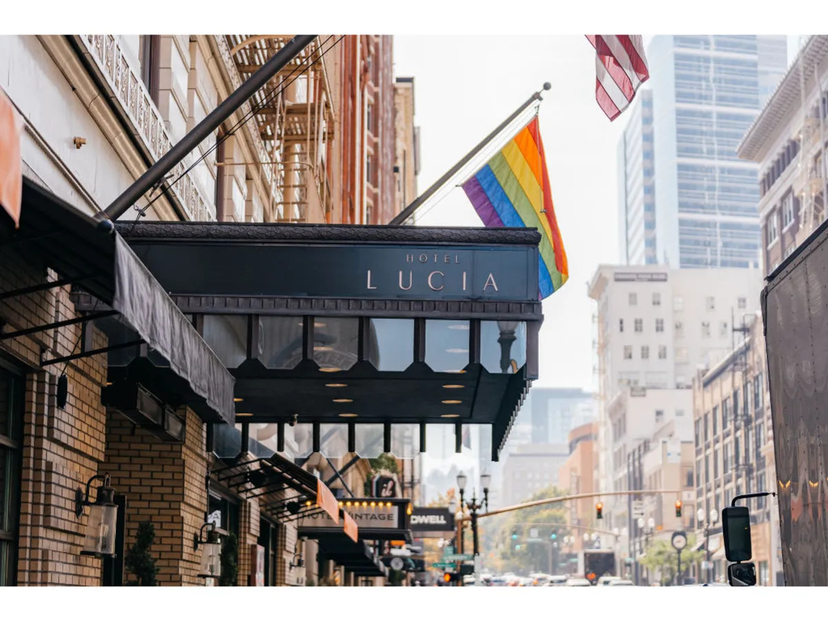 This exterior shot captures the Hotel Lucia entrance adorned with a rainbow flag and American flag, creating a vibrant and welcoming atmosphere. The hotel sign is prominently displayed, and the surrounding street hints at the lively urban environment just outside the doors.