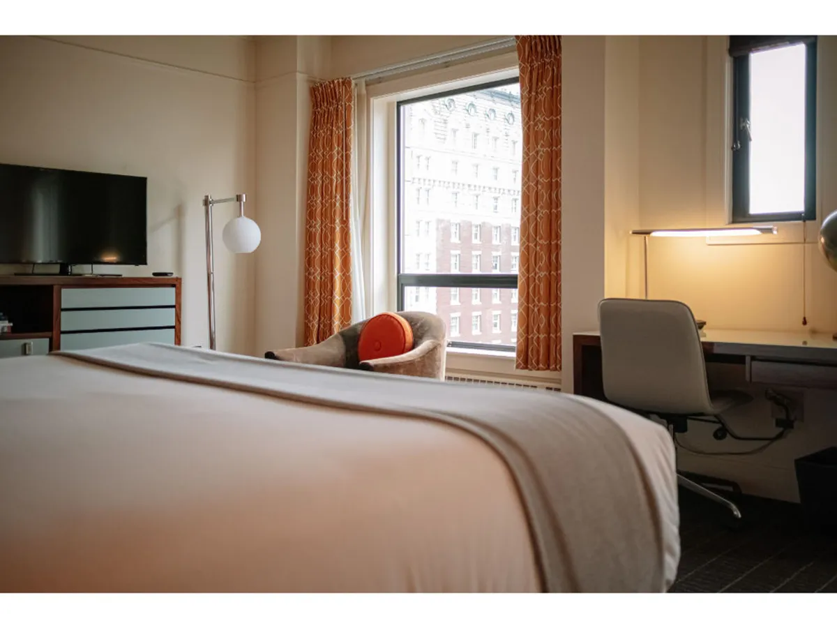 A guest room featuring a bed with a gray blanket and bright orange accent pillow, a window showcasing a city skyline, a desk with a chair and lamp for productivity, and a TV on a wooden dresser. The space feels stylish and comfortable, ideal for both relaxation and work.
