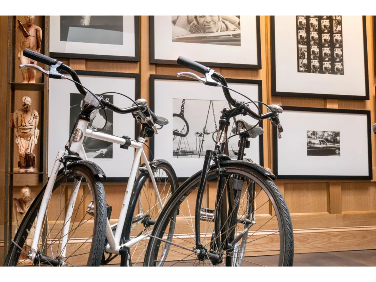 This image features two Shinola bicycles—one white and one black—against a backdrop of black and white framed photographs and stylish wooden wall paneling. A small sculpture adds artistic flair, and the overall setting feels dynamic and creative. The scene hints at adventure and exploration, with the bicycles front and center.