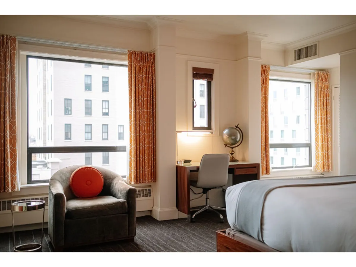 This image features a comfortable interior setting with a large window that offers views of the surrounding cityscape. The room includes an inviting armchair with a red cushion, a desk with a globe that hints at exploration and productivity, and a bed with crisp white linens. Natural daylight pours in, blurring the boundary between the inviting interior and the energetic city life just outside.