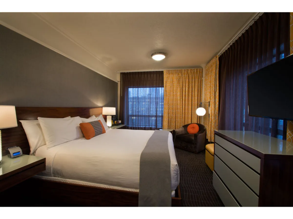A welcoming guest room with a plush bed adorned with decorative pillows, an armchair positioned near a large window offering a city view, and a flat-screen television on the wall. The natural light pouring in through the window highlights a cozy ambiance, suggesting relaxation after a day spent outdoors.