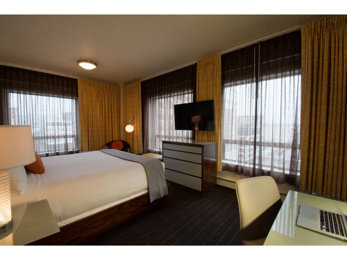 A modern guest room is shown with a comfortable bed, window curtains open to let in soft natural light, a television, and a dedicated desk. The atmosphere feels relaxed yet functional, with space for both restful downtime and productive work. The window suggests a connection to the outdoors and the urban environment beyond.