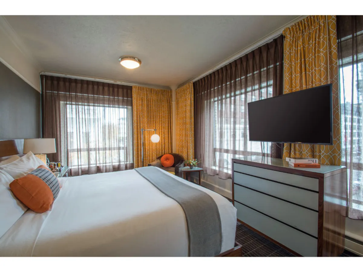 The image showcases a stylish hotel room in downtown Portland with a plush bed adorned with decorative pillows, large windows letting in streams of natural light through sheer curtains, a modern dresser, and a wall-mounted TV. The atmosphere is bright, fresh, and inviting, perfect for relaxation or unwinding after a day in the city.