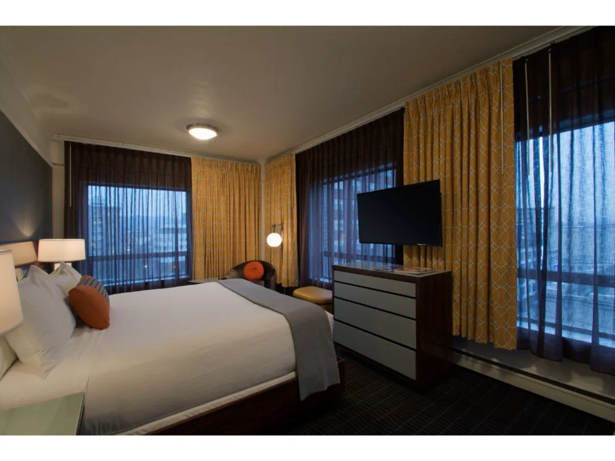 A bright, inviting bedroom featuring white linens, an orange accent pillow, a large television, and textured curtains. The layout suggests a restful environment, ideal for relaxing after a day spent exploring the city with a pet.