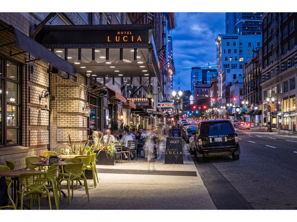 This exterior image highlights the Hotel Lucia sign, an inviting outdoor seating area filled with diners, illuminated street lights, and the lively energy of people socializing. The setting is urban and bustling, perfectly embodying the downtown atmosphere.