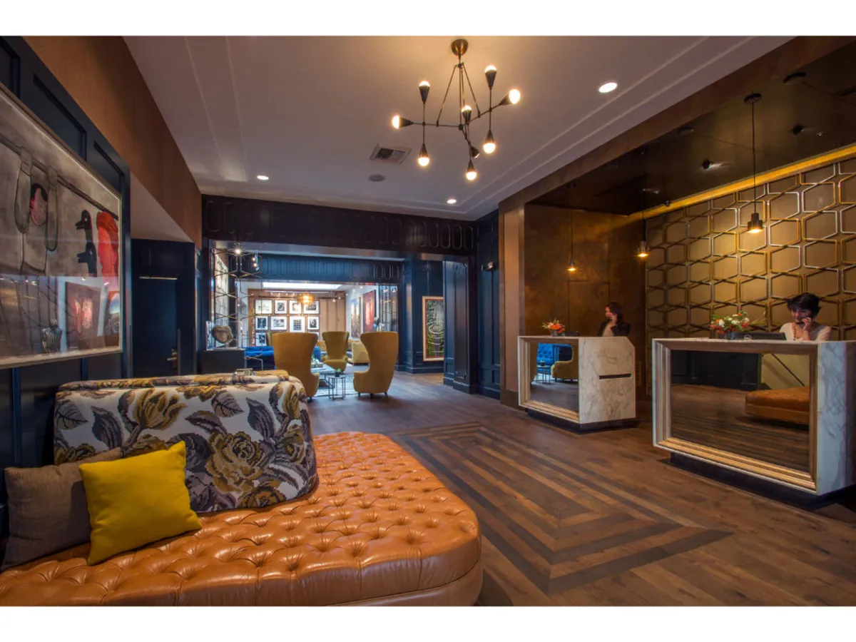 A contemporary lounge area with a tufted leather sofa, stylish reception desks, and modern lighting. The ambiance is welcoming, with guests visible either relaxing or approaching the check-in area. Unique artwork enhances the sophisticated hotel atmosphere.