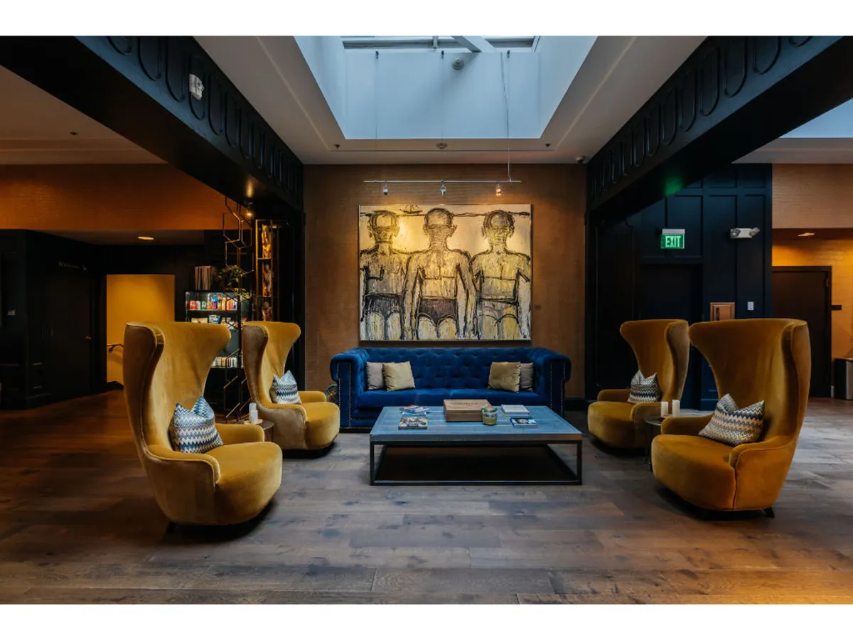A cozy, creative lounge featuring a blue sofa and yellow armchairs, complemented by artwork on the wall and a central coffee table. The space invites guests to socialize, relax, read, or enjoy art showcases in a comfortable, community-driven setting.